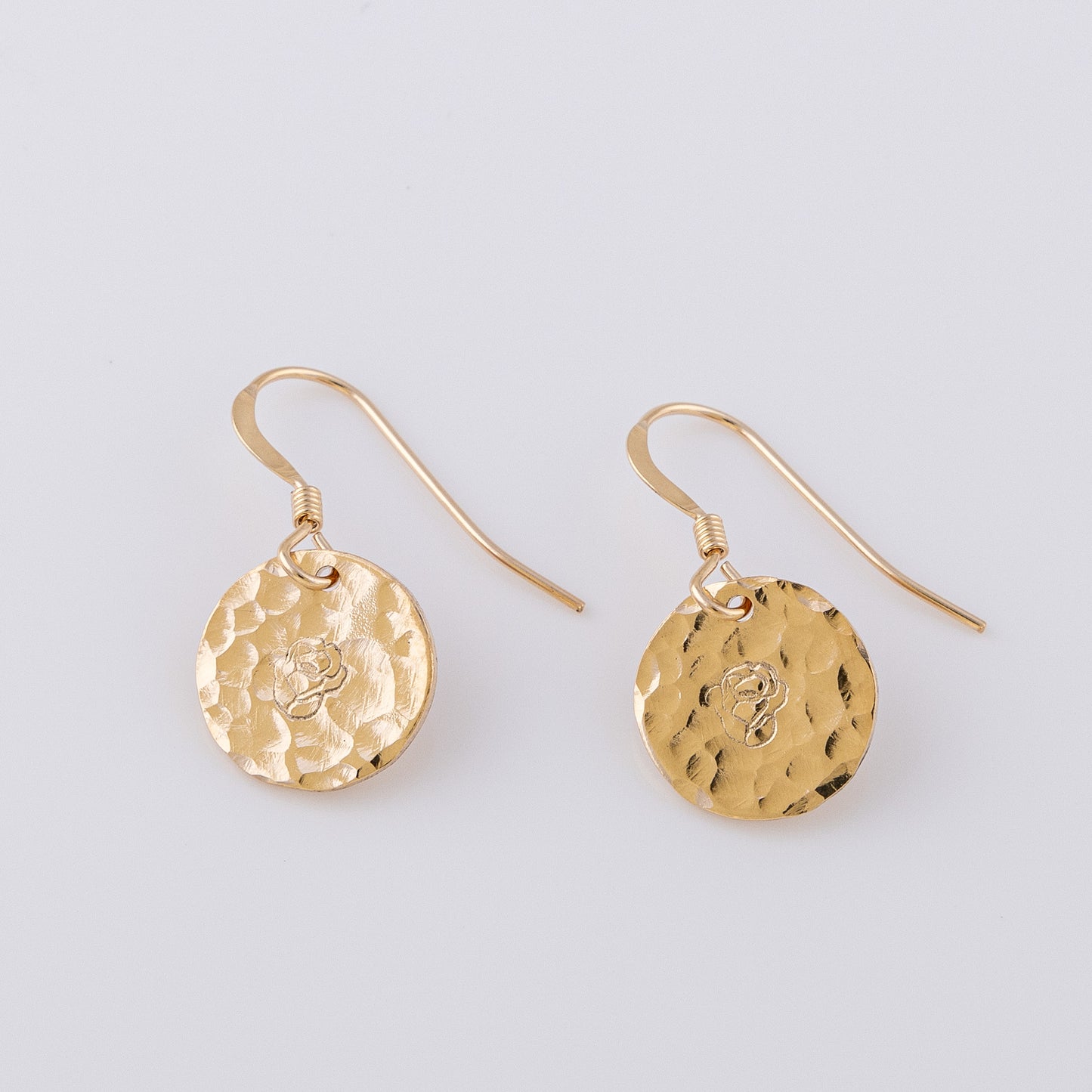 Two fish hook earrings. 14ct gold-fill, disc earrings with a hammered finish. The disc is hand stamped with a rose. One earring is facing the light, the textured finish catching teh light and creating a sparkle. The other faces away from the light.