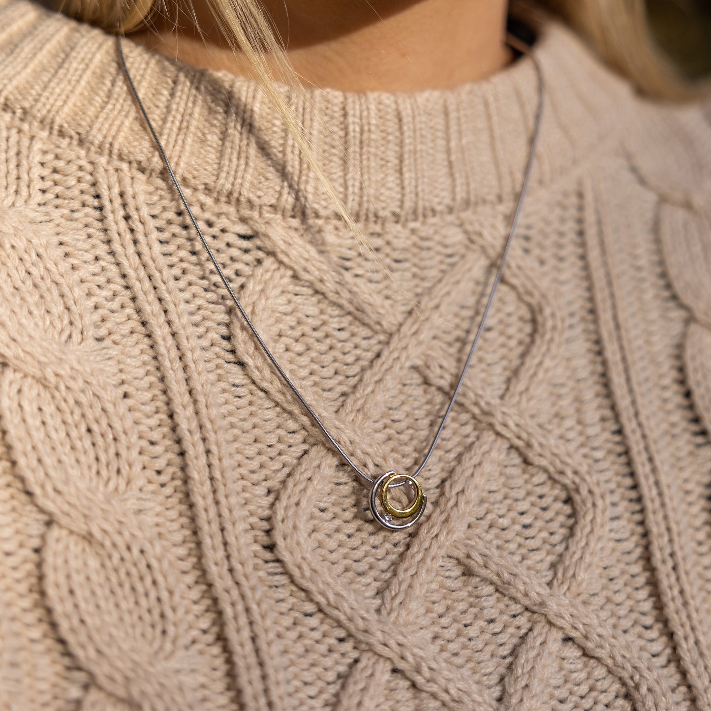 Gold vermeil and rhodium plated silver necklace with white cubic zirconia detail. The pendant has a circle design and sits on a rhodium plated silver, snake chain. The necklace is worn on top of a beige aran jumper with a round neck.