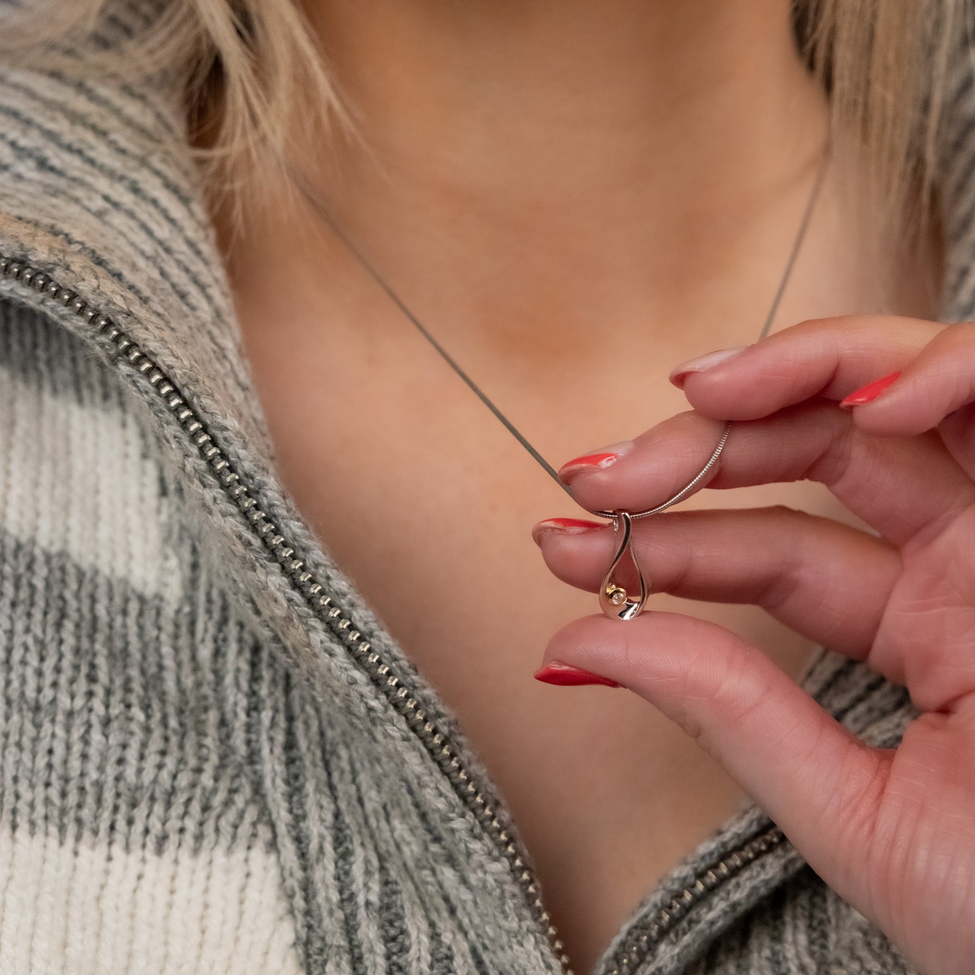 Shot of models chest, she is holding the pendant of the necklace shes wearing between hwe middle finger and thumb and showing it to the camera. The pendant is tear shaped is rhodium plated silver with gold vermeil detail and a single set diamond. The chain is a rhodium plated silver snake chain