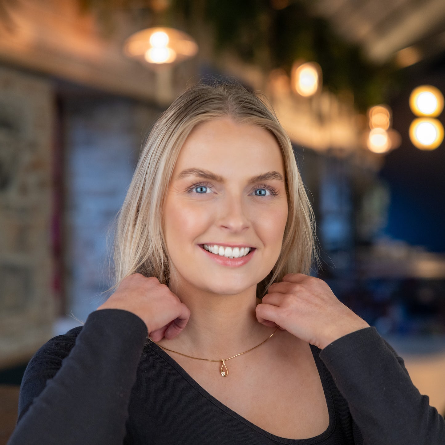 Model, a young blonde woman, smiling at the camera. She is touching a gold vermeil pendantr necklace she is wearing. The pendant is a tear drop shape with a set diamond detail, the chain is a gold vermeil snake chain. The model is wearing a long sleeve black top with an asymetrical neckline