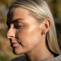 Model, a young blonde haired woman, outdoors wearings rhodium plated silver stud earrings. The earrings have a butterfly clasp and are tear drop shaped. They have a gold vermeil detail surrounding the one set diamond on the earring.