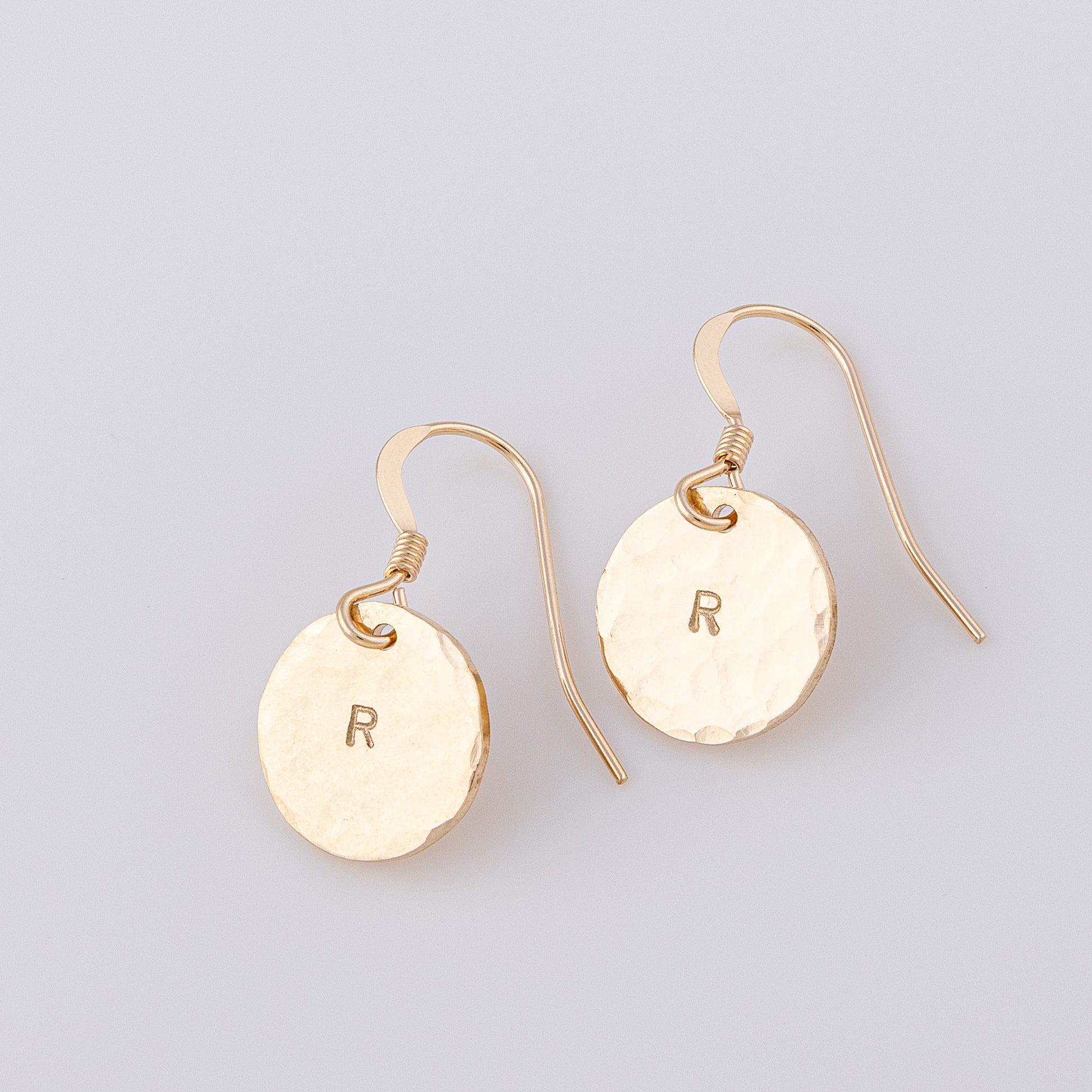 A side angle of two 14ct gold-fill disc earrings. These fish hook earrinsg are hand made and have a hammered finish with an initial stamped in the centre of the disc.