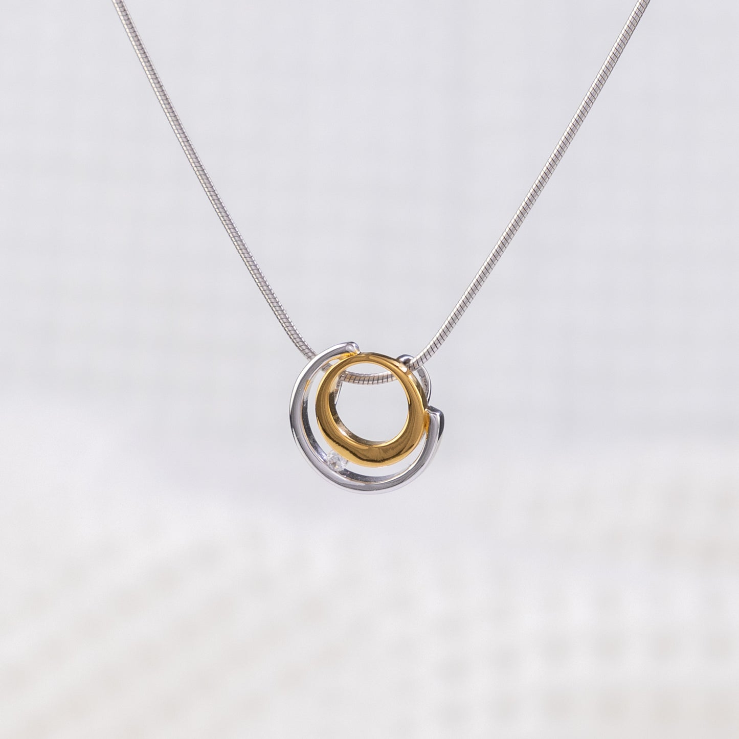 Rhodium plated silver and gold vermeil pendant with a white cubic zirconia detail. The pednant design is a circle design, surrounded by a semi circle with white cubic zirconia placed between the two. The pendant is on a rhodium plated silver, snake chain. The background of the image is white