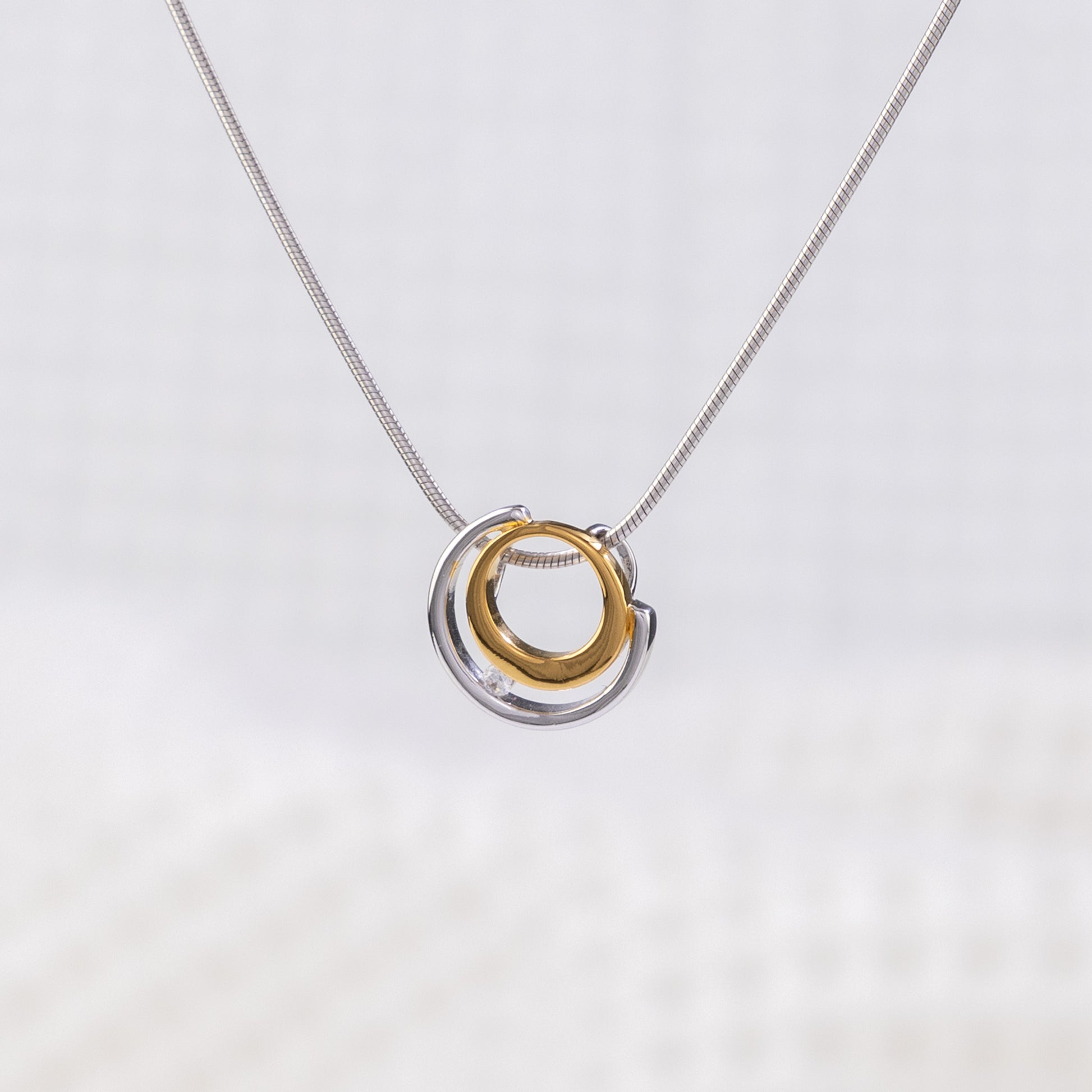 Rhodium plated silver and gold vermeil pendant with a white cubic zirconia detail. The pednant design is a circle design, surrounded by a semi circle with white cubic zirconia placed between the two. The pendant is on a rhodium plated silver, snake chain. The background of the image is white