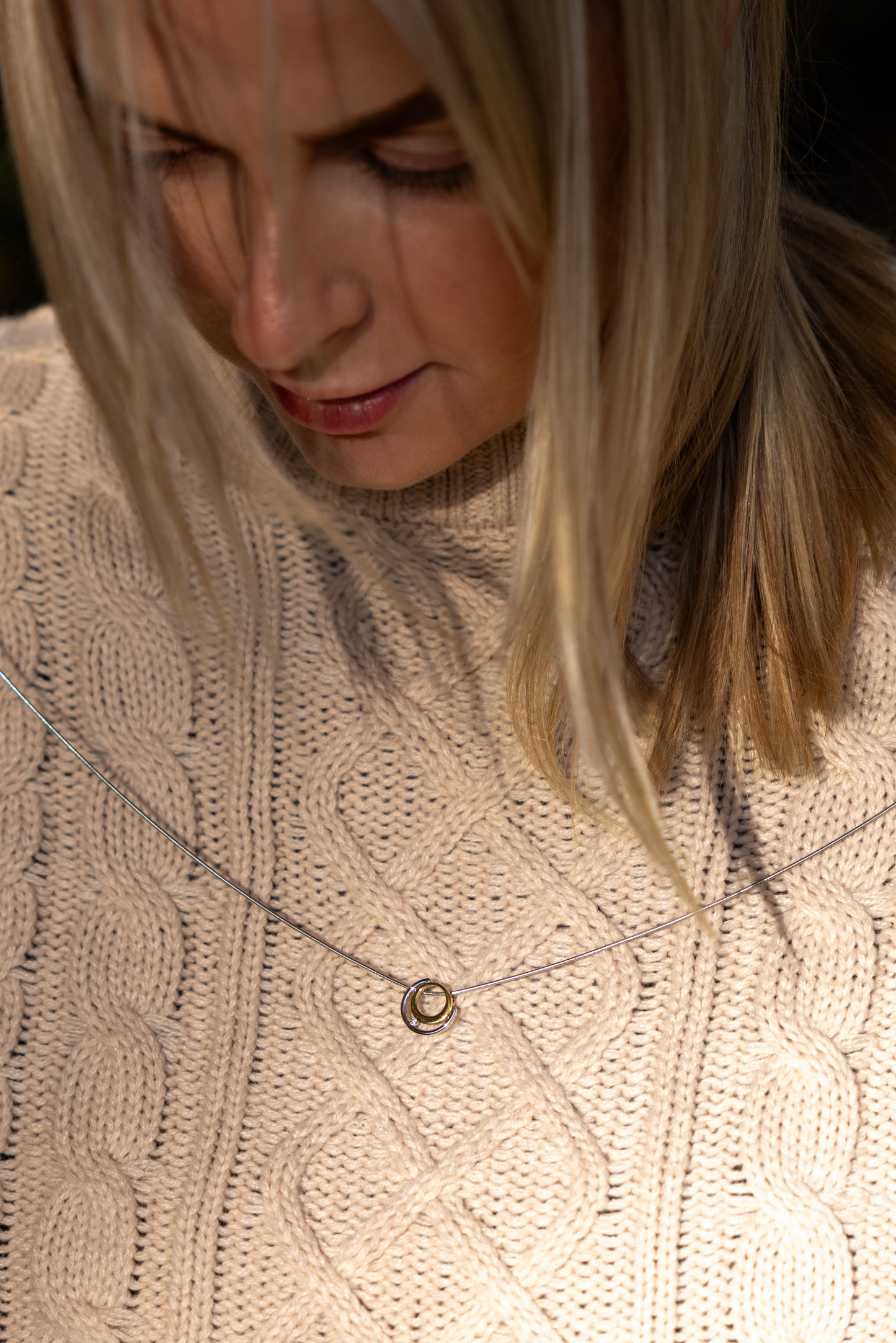 Model, a young blonde woman putting on a gold vermeil and rhodium plated silver necklace with white cubic zirconia detail. The pendant is a circle design and sits a top a rhodium plated silver chain. The model has a beige aran jumper on with a round neck and is tilting her head down to put tha necklace on.