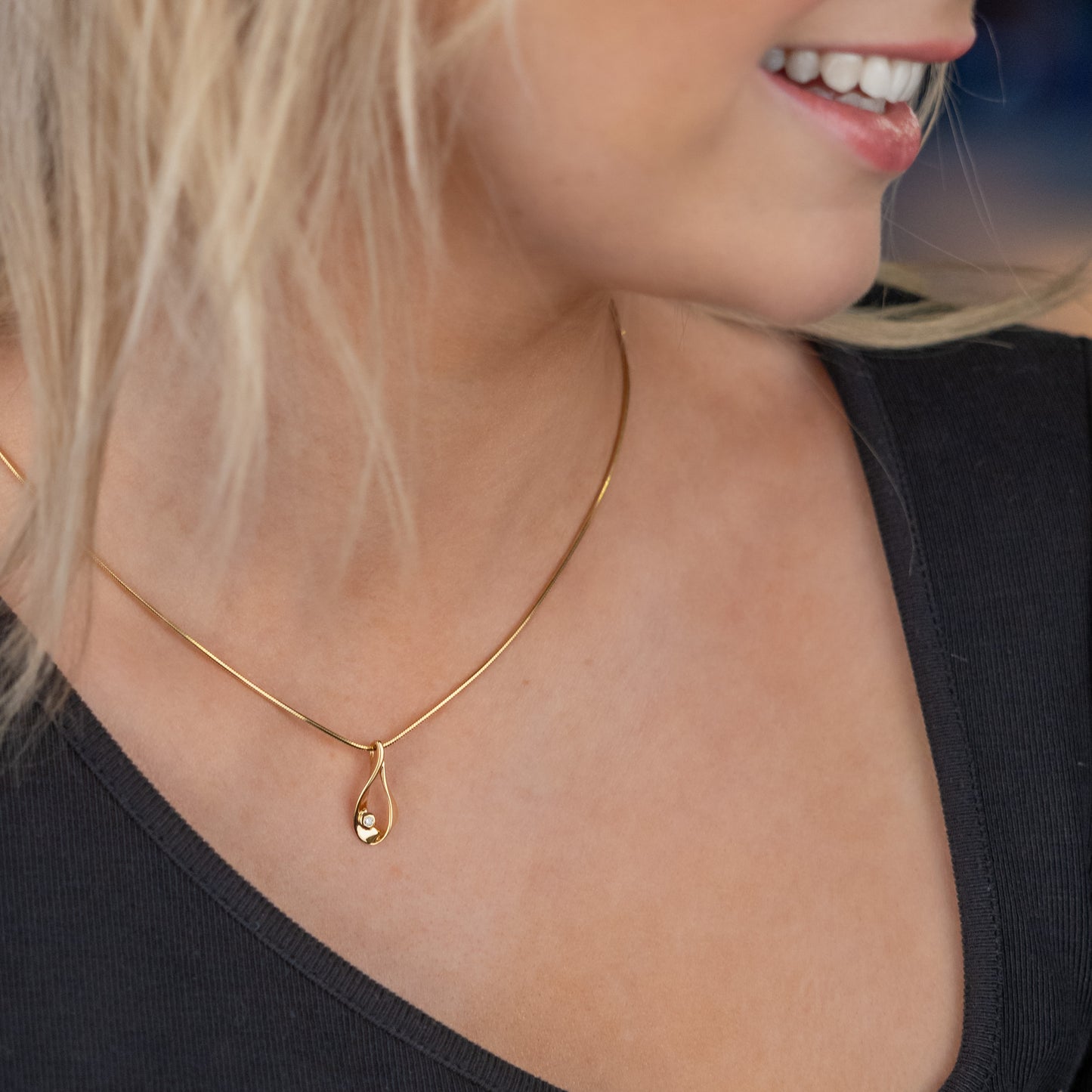 A gold vermeil pendant necklace on a young woman wearing a asymetrical neckline black top. The pendant is a tear drop shape with a set diamond detail. The chain is a gold vermeil snake chain