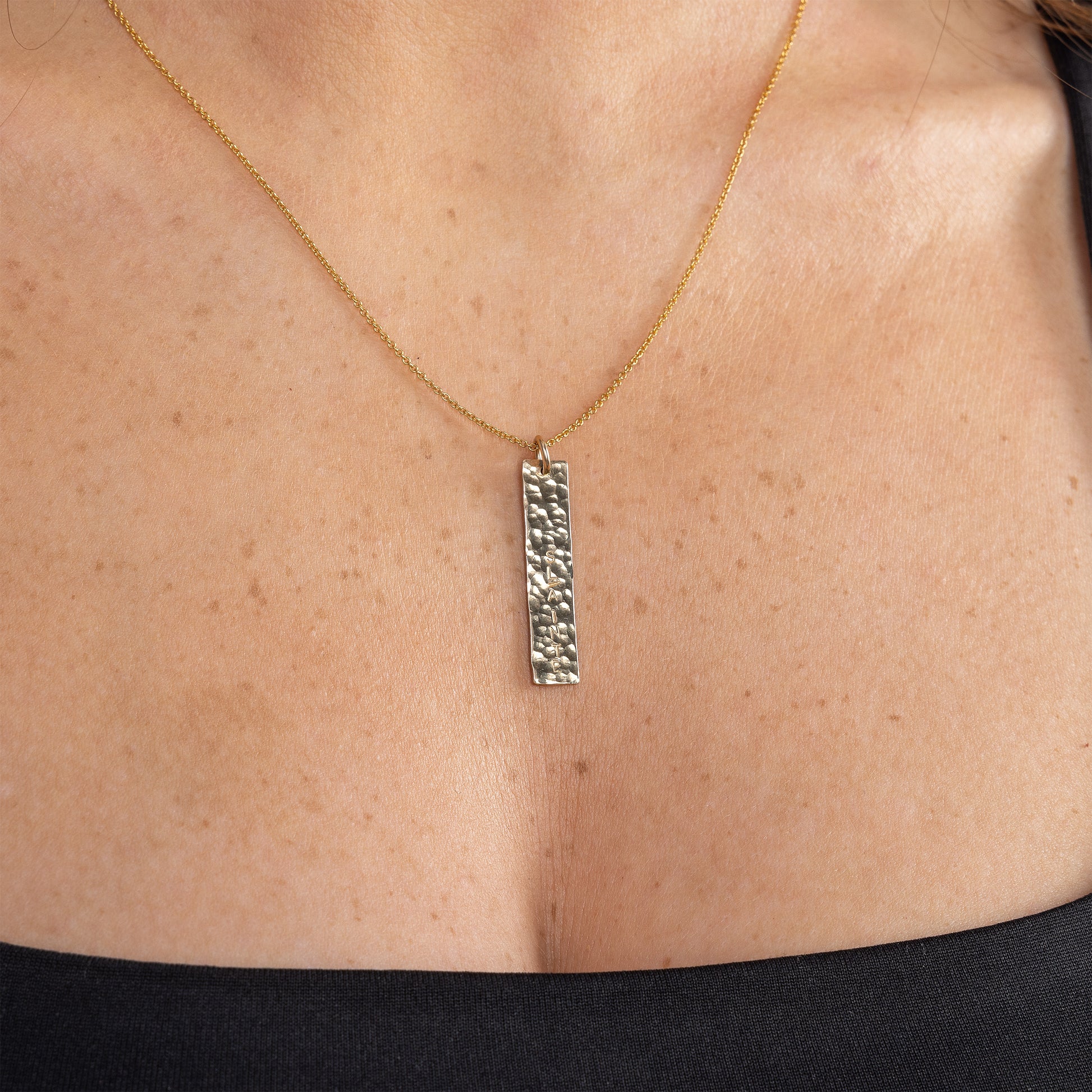 Model wearing a 14ct gold filled hammered bar pendant necklace. The pendant is personalised with a custom word of your choice/ a name handstamped. The bar pendant is a textured finish that sparkles in the light. 