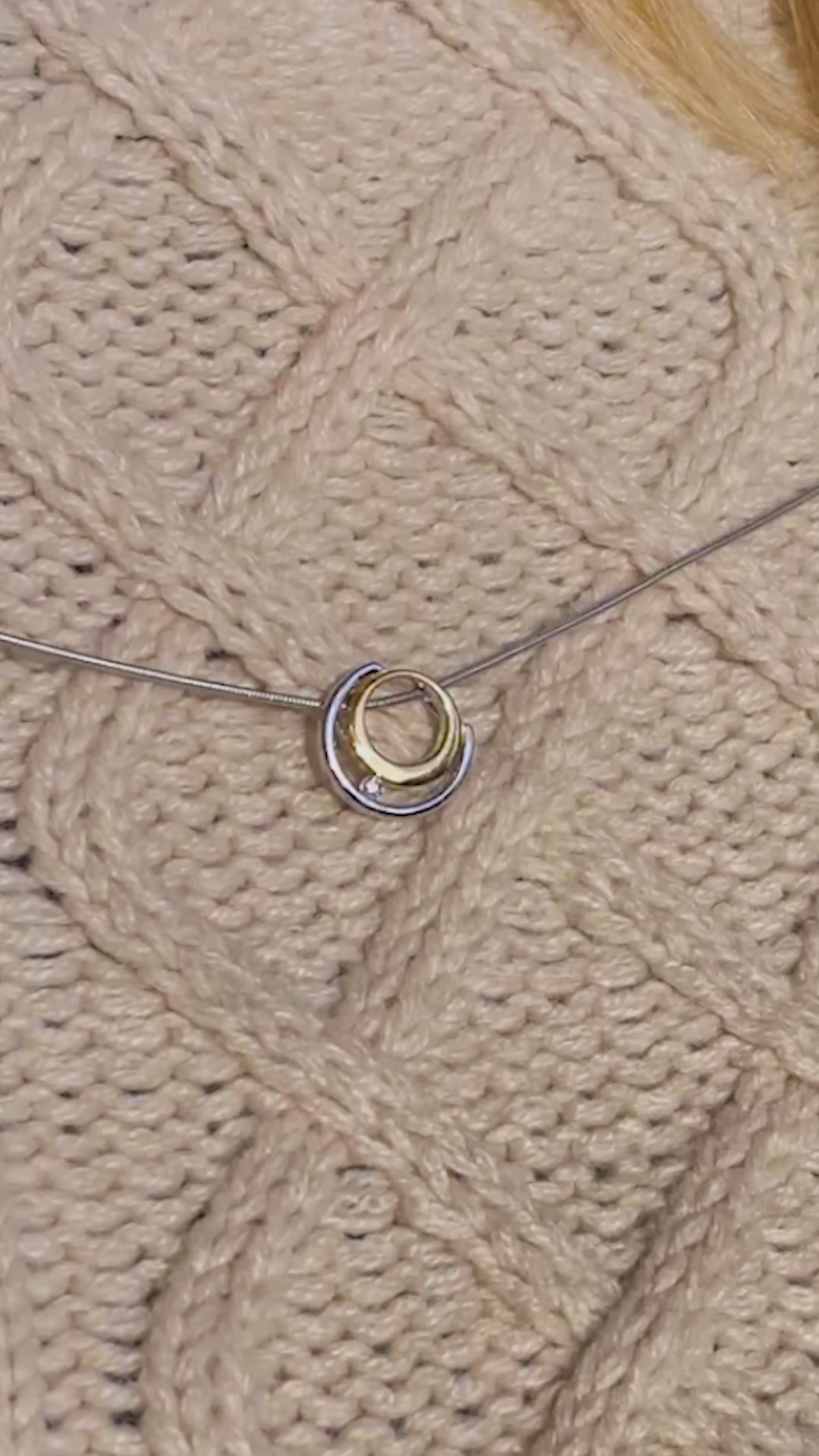  Video of a gold vermeil and rhodium plated silver necklace with white cubic zirconia detail worn by a young blonde haired woman. The pendant has a circle design and sits on a rhodium plated silver, snake chain. The necklace is worn on top of a beige aran jumper with a round neck. The video is filmed outside with the sun shining on the models face