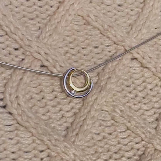  Video of a gold vermeil and rhodium plated silver necklace with white cubic zirconia detail worn by a young blonde haired woman. The pendant has a circle design and sits on a rhodium plated silver, snake chain. The necklace is worn on top of a beige aran jumper with a round neck. The video is filmed outside with the sun shining on the models face